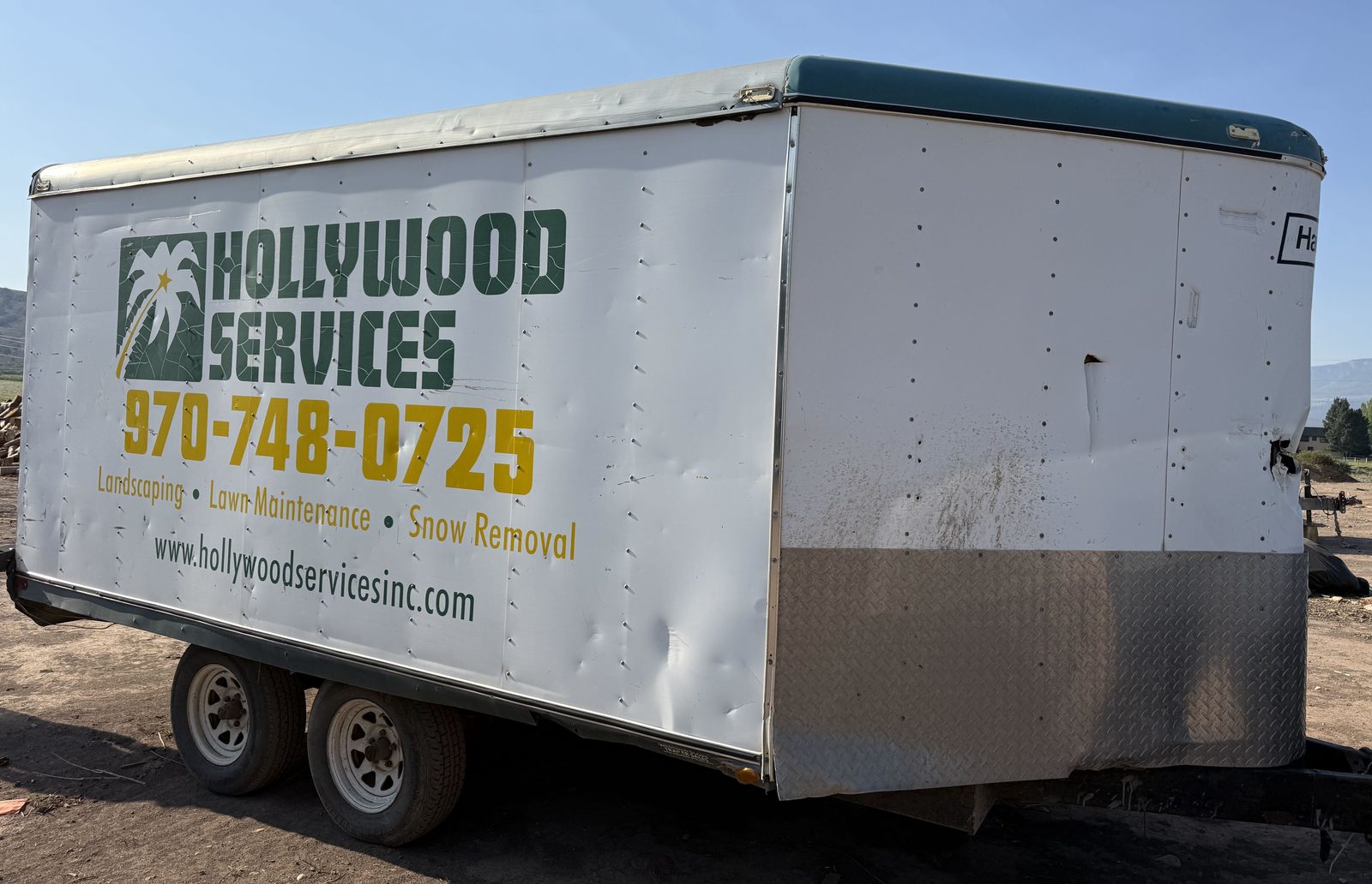 A Hollywood Services equipment trailer in its original green and gold livery, listing landscaping, lawn maintenance, and snow removal.